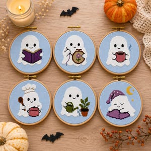 May include: Six circular wooden embroidery hoops featuring cross-stitch designs of white ghosts on light blue fabric. The ghosts are depicted reading a book, sewing, drinking from a mug, cooking with a pot, watering a plant, and sleeping on a pillow.