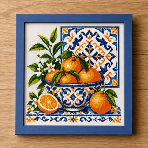 May include: A framed cross-stitch embroidery depicting a blue and white patterned bowl filled with bright orange citrus fruits and green leaves. The design includes a halved orange, white blossoms, and a decorative tile-inspired background.