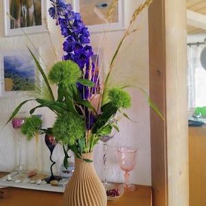 Könnte beinhalten: Eine hellbeige Vase mit vertikaler Rippenstruktur enthält ein Blumenarrangement mit hohen, leuchtend violetten Ritterspornstielen, hellgrünen Bartnelkenblüten und dünnen, feinen Halmen aus getrocknetem Ziergras.