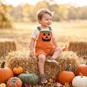 May include: A pair of orange corduroy overalls featuring a black jack-o'-lantern face appliqué on the chest, worn over a short-sleeved cream-colored t-shirt. The overalls have green shoulder straps and are paired with brown sneakers.