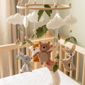 May include: A nursery mobile featuring a circular wooden frame with hanging felt elements including white clouds, green leaves, and stuffed animal figures of a gray rabbit, an orange fox, a brown bear, and a tan deer, all suspended by white strings.