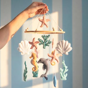 Può includere: Mobile per bambini fatto a mano con creature marine in feltro. Il mobile include coralli, stelle marine, conchiglie, un cavalluccio marino e un delfino in tonalità rosa, verde acqua e grigio, sospesi da un anello di legno.