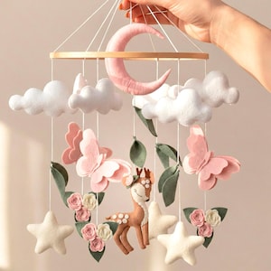 May include: A nursery mobile with a wooden frame, a pink crescent moon, white clouds, butterflies, stars, and a deer. The mobile is adorned with felt flowers and leaves, creating a whimsical decoration for a baby's room.