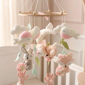 Puede incluir: Móvil para bebé con animales y nubes de fieltro. Incluye un conejito, un zorro, pájaros y nubes en colores rosa, blanco y gris suaves. Cuelga de un aro de madera con cuentas de madera.