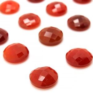 May include: A collection of 12 red carnelian gemstone cabochons, each with a faceted surface. The cabochons are arranged in a scattered pattern on a white background.