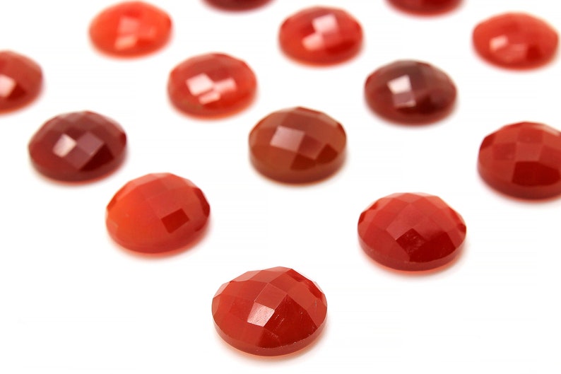 May include: A collection of 12 red carnelian gemstone cabochons, each with a faceted surface. The cabochons are arranged in a scattered pattern on a white background.