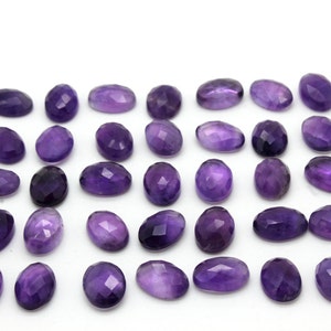 May include: A collection of 30 oval-shaped amethyst gemstones, each with a faceted cut. The stones are a deep purple color and are arranged in rows on a white background.
