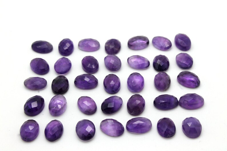 May include: A collection of 30 oval-shaped amethyst gemstones, each with a faceted cut. The stones are a deep purple color and are arranged in rows on a white background.