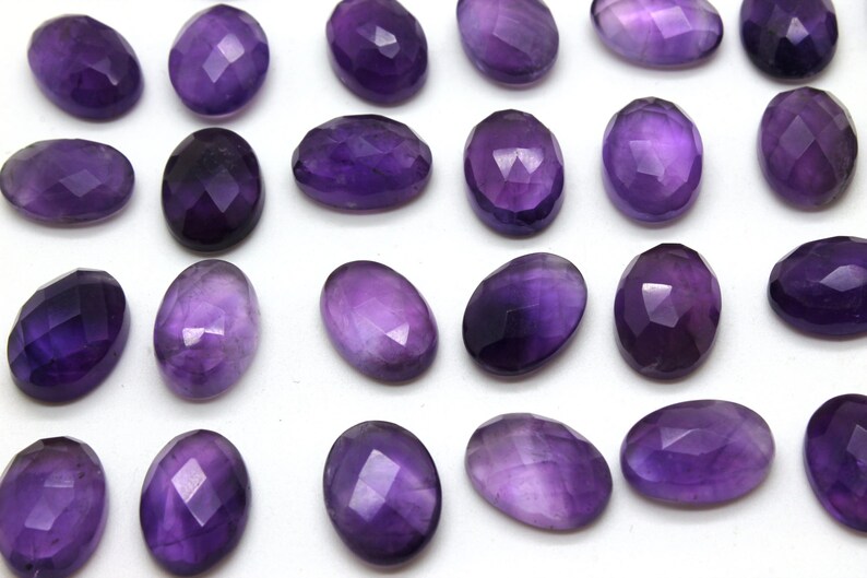 May include: A collection of 20 faceted amethyst gemstones, each with a smooth, oval shape. The stones are a deep purple color and have a glossy finish.