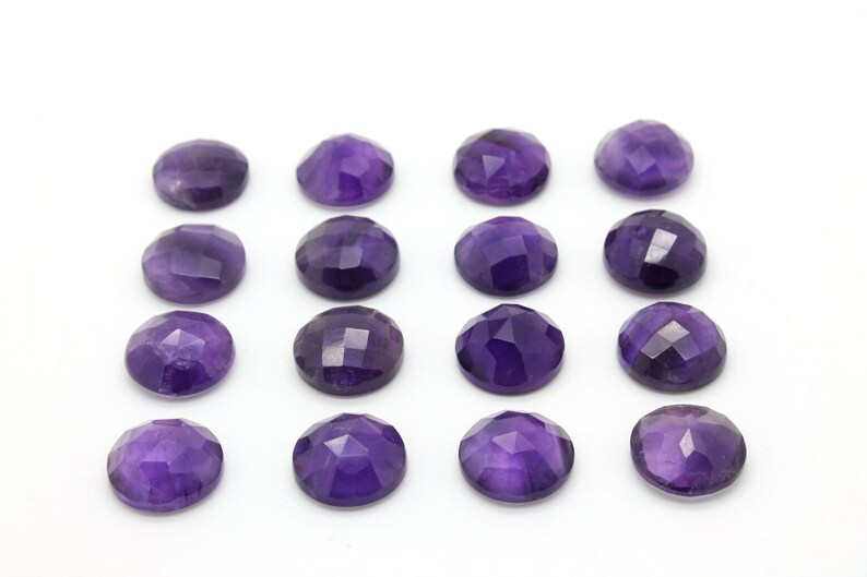 May include: A collection of 16 round, faceted amethyst gemstones. The stones are a deep purple color and are arranged in four rows of four.