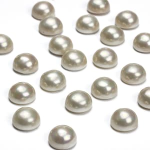 Puede incluir: Un conjunto de perlas semiredondas de color blanco roto, dispersas sobre una superficie blanca. Cada perla tiene una superficie lisa y abovedada con un sutil brillo iridiscente. Las perlas son uniformes en tamaño y forma, probablemente para joyería.