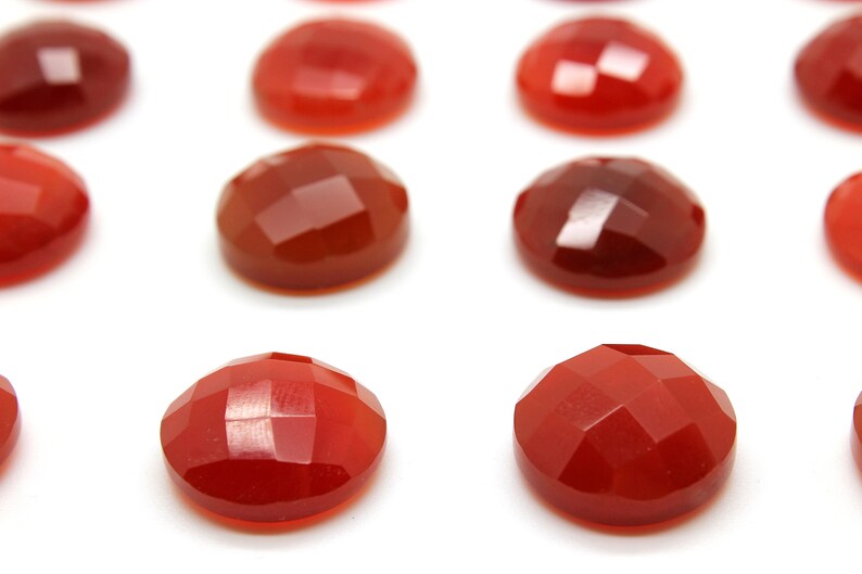 May include: A collection of 12 red carnelian gemstones, each with a faceted, rounded shape. The stones are arranged in rows on a white surface.