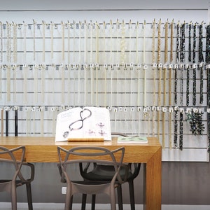 May include: A white wall with rows of necklaces hanging on hooks. The necklaces are made of various materials, including pearls, beads, and stones. There is a wooden table with two chairs in front of the wall. A book is open on the table.