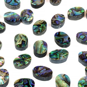 Puede incluir: Una colección de cabujones ovalados de concha de abulón en varios tonos de verde, azul y morado. Los cabujones son iridiscentes y tienen un aspecto natural y orgánico.