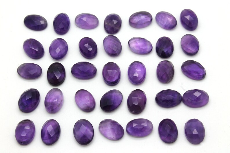 May include: Thirty-six oval-shaped amethyst gemstones, each with a faceted cut, arranged in rows on a white background.