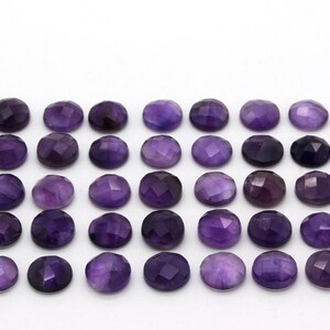 May include: A collection of 40 round, faceted amethyst gemstones. The stones are a deep purple color and are arranged in rows on a white background.