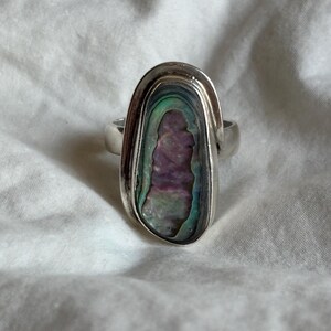 May include: A silver ring featuring an oval abalone shell inlay with iridescent purple, green, and blue hues. The ring has a polished silver band and a bezel setting that frames the shell.