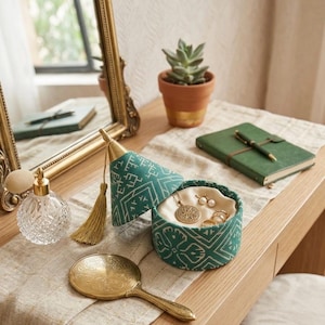Peut inclure: Une coiffeuse avec un miroir doré, un flacon de parfum en cristal et une boîte à bijoux verte à motifs. La boîte est ouverte, révélant des bijoux. Un miroir à main doré et une petite plante succulente en pot sont également présents.