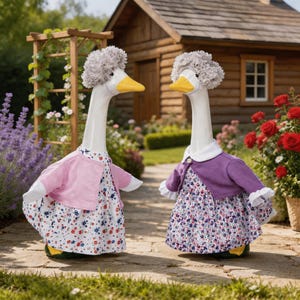 May include: Two white goose statues wearing gray yarn wigs, floral dresses, and cardigans. One goose wears a light pink cardigan over a white dress with red and blue flowers. The other wears a purple cardigan over a white dress with blue and red flowers.