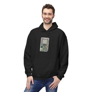 Puede incluir: Sudadera con capucha negra con un gráfico detallado de una consola de videojuegos portátil vintage. El gráfico incluye la pantalla, los botones y los componentes internos. La sudadera tiene un bolsillo delantero y una capucha con cordón.