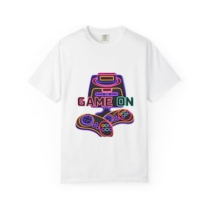 May include: White t-shirt featuring a neon-style graphic of a retro video game console and two controllers. The phrase "GAME ON" is displayed below the console in a colorful font. The design has a vibrant, retro aesthetic.