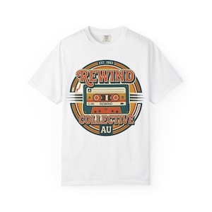 May include: White t-shirt featuring a retro design with the words "REWIND" and "COLLECTIVE AU" surrounding a cassette tape graphic. The design includes orange, green, and brown colors, with the text "EST. 1983" at the top.