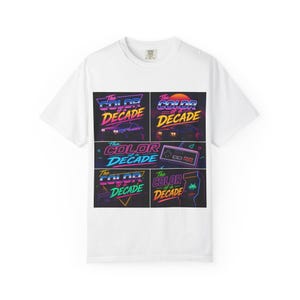 May include: White t-shirt featuring a retro design with the text "The Color of a Decade" in a vibrant, neon color scheme. The design includes images of a car, a sunset, a video game controller, and arcade game graphics.