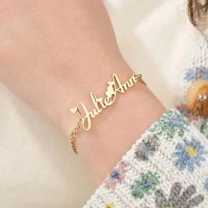 Puede incluir: Una pulsera dorada con los nombres "Julie Ann" en escritura cursiva. La pulsera se lleva en la muñeca y tiene una cadena fina. El diseño incluye un corazón y un pequeño elemento decorativo entre los nombres.