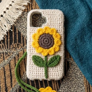 Puede incluir: Una funda de teléfono de ganchillo color crema con un girasol grande y tridimensional de pétalos amarillos y centro marrón oscuro. Un tallo verde con dos hojas se extiende hacia abajo y un dije de girasol a juego cuelga de un cordón verde.