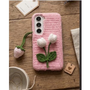 May include: A pink crocheted phone case featuring two white tulip flowers with green stems and leaves attached to the center. A small, light pink crocheted strawberry charm hangs from a green cord on the side of the rectangular protective cover.