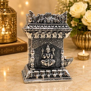 May include: A silver-toned metal oil lamp featuring an intricate relief of a seated deity on the front panel. The rectangular structure has ornate decorative borders, a tiered top, and a small protruding bowl on the side for a wick.