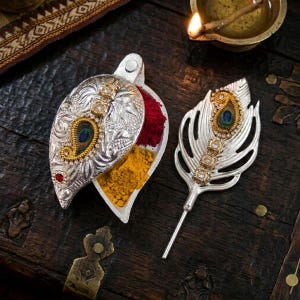 Puede incluir: Un recipiente plateado en forma de hoja con intrincados patrones en relieve, que contiene polvos rojos y amarillos, junto con un aplicador a juego en forma de pluma con un motivo central de ojo de pavo real y pequeñas piedras claras.