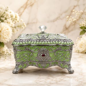 May include: A rectangular jewelry box featuring a light green enameled surface with intricate silver-toned metal filigree overlays. The lid is topped with a clear faceted crystal knob, and the front displays a central oval red gemstone accent.