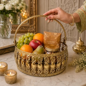 May include: A round gold metal basket with a decorative filigree pattern and a swing handle, filled with green grapes, two oranges, a red apple, a green pear, a jar of nuts, and a brown package labeled Luxury Dry Fruits.