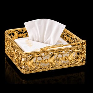 May include: A square gold-colored metal napkin holder featuring an intricate filigree pattern with circular motifs and floral accents. The holder contains a stack of white paper napkins, each decorated with a subtle gold feather print design.