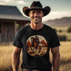 Könnte beinhalten: Schwarzes T-Shirt mit einem Aufdruck eines Cowboys auf einem Pferd, begleitet von einem Hund, in einer Wüstenlandschaft. Die Person, die das T-Shirt trägt, lächelt und trägt einen Cowboyhut. Im Hintergrund ist ein Holzgebäude und ein Feld zu sehen.