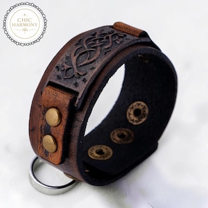 Puede incluir: Una pulsera de cuero marrón con un diseño de nudo celta. La pulsera presenta una banda ancha con un patrón en relieve detallado, asegurada con broches de color latón y un anillo plateado. El texto "CHIC HARMONY" está en un logotipo circular.