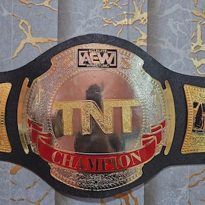 May include: A wrestling championship belt with a black leather strap and a large, ornate silver and gold buckle. The buckle features the text "TNT CHAMPION" in red, with the AEW Wrestling logo. The belt is designed with intricate details.