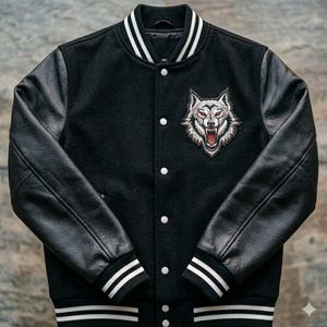 Op de afbeelding: Zwarte varsity jas met zwarte leren mouwen en een wit en zwart gestreepte kraag en manchetten. De jas heeft een wolfskop patch met rode ogen op de borst. De jas heeft witte knopen.