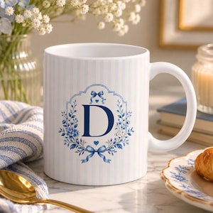 Könnte beinhalten: Ein weißer Keramikbecher mit hellblauem vertikalem Streifenmuster und einem zentralen dunkelblauen Buchstaben D, der von einem dekorativen Blumenkranz mit blauen Schleifen umrahmt ist. Der Becher hat eine glatte Oberfläche und einen geschwungenen Henkel.