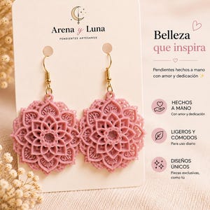 Puede incluir: Un par de pendientes rosas con forma de mandala y ganchos dorados. Los pendientes se exhiben en una tarjeta blanca con el texto "Arena y Luna" y "PENDIENTES ARTESANOS". La tarjeta también presenta el texto "Belleza que inspira".
