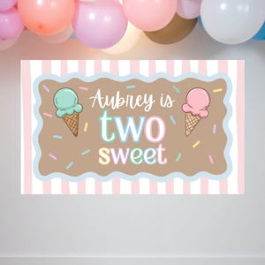 May include: A rectangular sign with the text "Aubrey is two sweet" in colourful lettering, featuring ice cream cone illustrations. The background has pink and white stripes, with a light blue border and sprinkles.