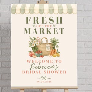 Op de afbeelding: Een bord op een ezel met de tekst "FRESH OFF THE MARKET" boven een aquarel illustratie van een mand met producten. Het bord luidt "WELCOME TO Rebecca's BRIDAL SHOWER" en de datum "06.20.2026". De bovenkant heeft een groen en wit gestreepte luifel.