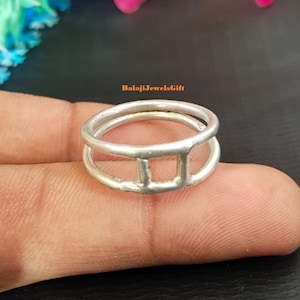 Può includere: Un anello color argento caratterizzato da un design a doppia fascia collegato da una barra verticale. L'anello presenta una finitura metallica liscia e una struttura semplice e aperta. Il testo BalajiJewelsGift è visibile in arancione sopra l'anello.