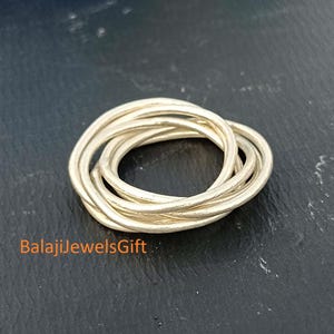 Può includere: Un anello color argento composto da molteplici fasce sottili intrecciate che creano una struttura circolare a strati. La superficie metallica ha una finitura opaca. Il testo BalajiJewelsGift è stampato in arancione nell'angolo in basso a sinistra.