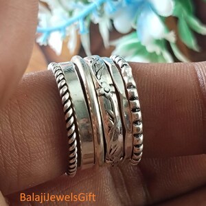 May include: A set of five stacked silver rings featuring varied textures including a twisted rope band, a smooth polished band, a plain thin band, a floral engraved band, and a beaded band. The text BalajiJewelsGift is visible at the bottom.