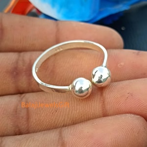 Puede incluir: Un anillo abierto de color plateado que presenta dos cuentas esféricas pulidas en cada extremo de la banda. La superficie metálica es lisa y reflectante, con el texto BalajiJewelsGift impreso en naranja en el lado inferior izquierdo.