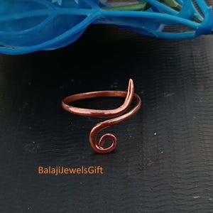 May include: A copper-coloured ring featuring an open, bypass-style band with one end terminating in a simple curve and the other end forming a small, tight spiral. The metal surface has a smooth, polished finish with a warm, reddish-brown metallic tone.