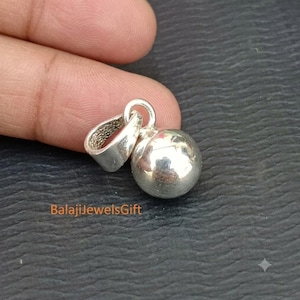 Könnte beinhalten: Ein polierter, kugelförmiger Anhänger aus Silber mit einem glatten, runden Design, das an einer stabilen, texturierten Öse befestigt ist. Die Metalloberfläche reflektiert das Licht und erzeugt helle Glanzlichter auf der Kugel. Der Text BalajiJewelsGift ist sichtbar.