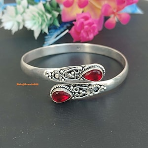 Pode incluir: Uma pulseira aberta de tom prateado com duas pedras vermelhas em forma de gota nas extremidades. O trabalho em metal ao redor de cada pedra exibe padrões intrincados de filigrana e texturas de contas. O texto BalajiJewelsGift é visível.